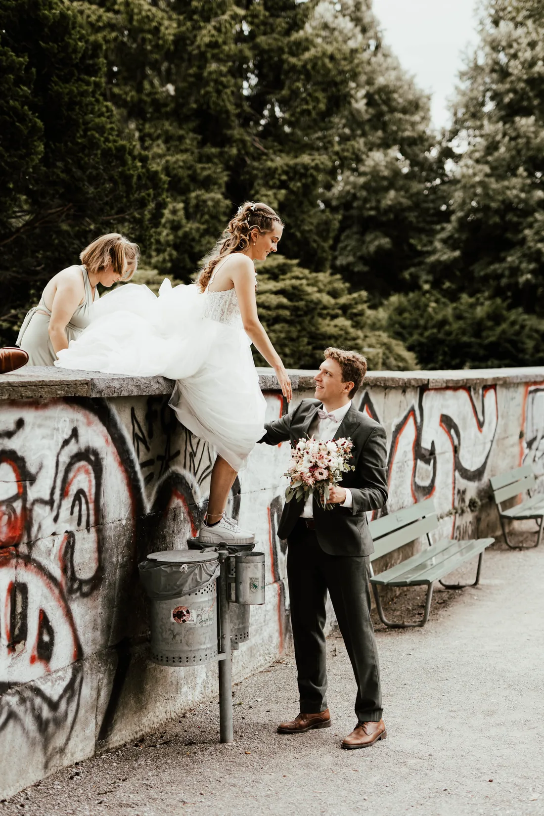 Braut sitzt auf einer Mauer mit Graffiti, der Bräutigam steht davor