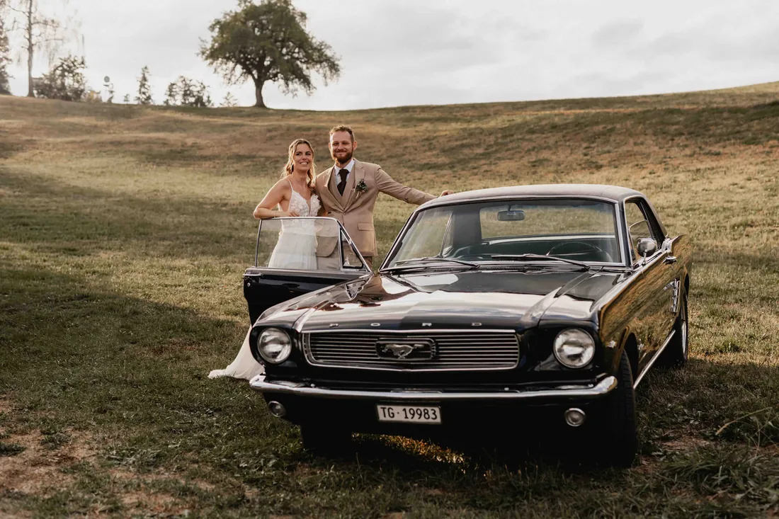 Brautpaar neben einem schwarzen Oldtimer auf einer Wiese