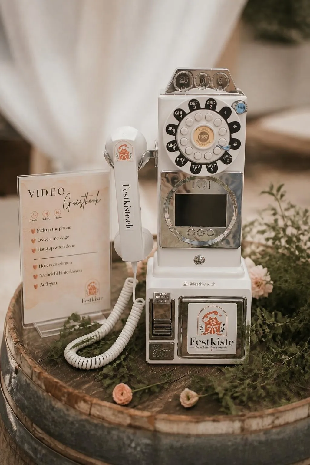 Video-Telefon von Festkiste: weisses Retro-Münztelefon mit Bildschirm, daneben eine Anleitung «Video Guestbook» auf einem Holzfass
